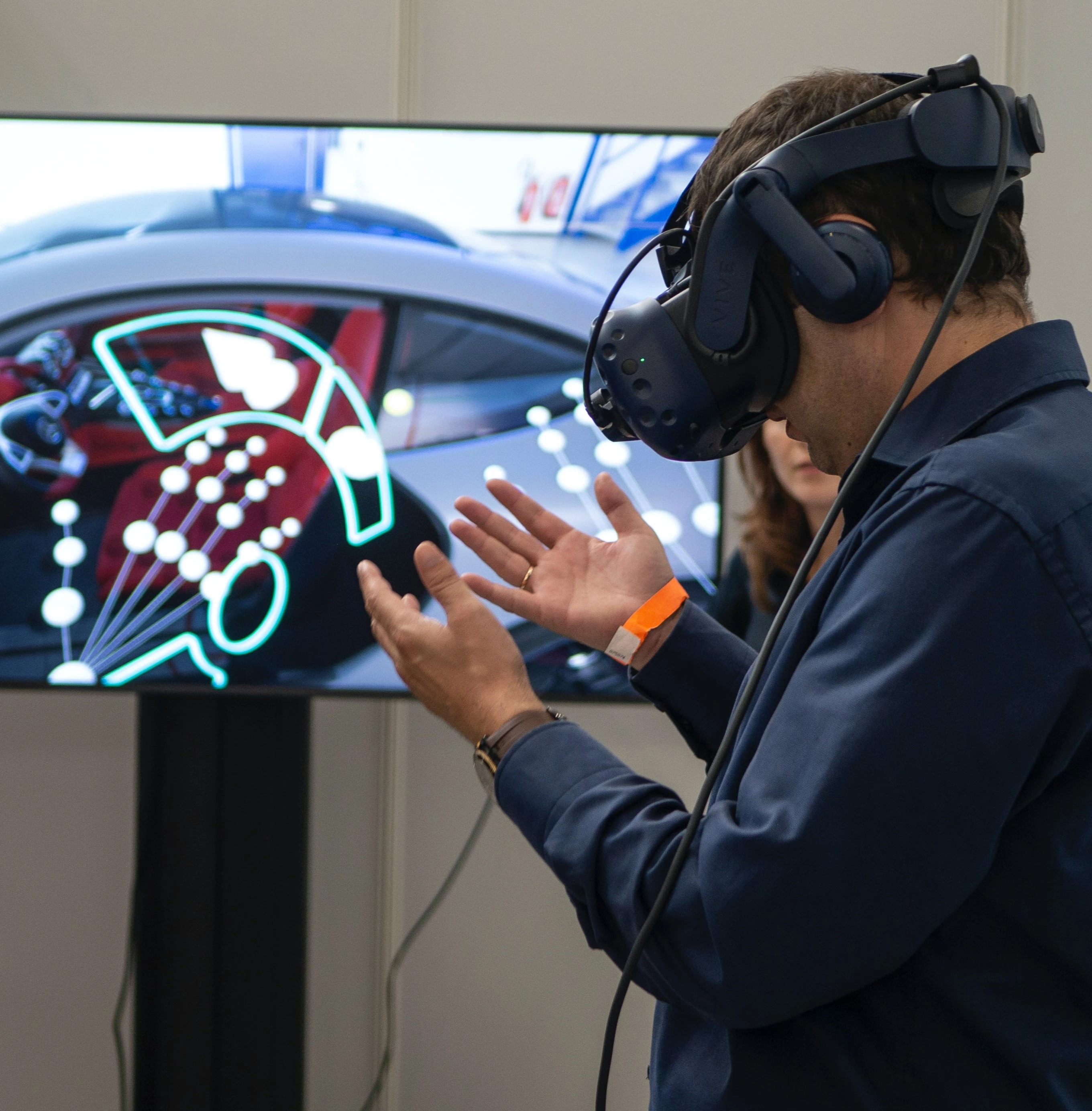 Man in a VR headset looking down at his hands. Screen behind him depicts what he is seeing- a VR simulation that allows you to explore a car model.